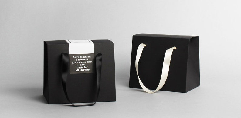 Op de afbeelding: Twee zwarte cadeauzakken met handvatten. E&eacute;n tas heeft een wit label met de tekst "Liefde begint in een moment, groeit met de tijd en duurt voor altijd".