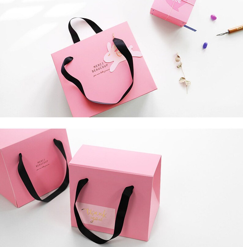 May include: Two pink gift bags with black ribbon handles. One bag has a bunny cutout and the text "Merci beaucoup" printed on it. The other bag has the text "Thank you" printed on it.