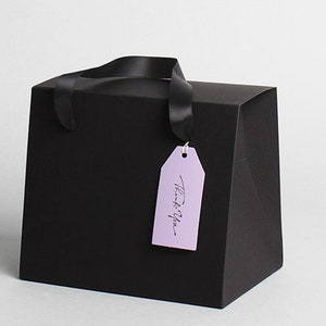 Op de afbeelding: Zwarte cadeauzak met een zwarte lintgreep en een lavendelkleurig label met de tekst "Bedankt".