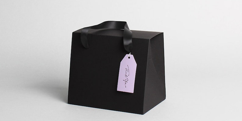 Op de afbeelding: Zwarte cadeauzak met een zwarte lintgreep en een lavendelkleurig label met de tekst "Bedankt".