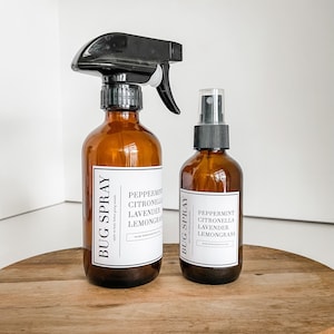 Op de afbeelding: Twee amberkleurige glazen flessen met zwarte spuitkoppen en witte etiketten. De etiketten lezen "BUG SPRAY" en bevatten ingrediënten zoals pepermunt, citroengras, lavendel en citroengras.