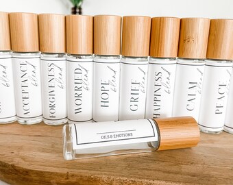 Puede incluir: Un conjunto de diez botellas de vidrio con rodillo, tapas de bambú y etiquetas blancas. Las etiquetas tienen las palabras "blend" y varias emociones escritas en ellas, como "acceptance", "forgiveness", "worried", "hope", "grief", "happiness", "calm" y "peace". Las botellas están dispuestas en una fila sobre una superficie de madera. El texto "OILS & EMOTIONS" está impreso en la etiqueta de una de las botellas.