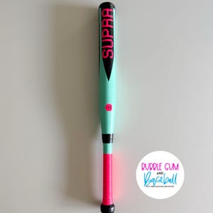 May include: A mint green and pink baseball bat with the word "SUPRA" in pink lettering. The bat has a black handle and a pink grip. The number "10" is printed in pink. The bat is on a white background.
