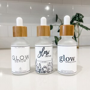 May include: Three glass bottles with bamboo lids and white labels. The labels have the text "glow serum" in black lettering. The middle bottle has a floral design on the label. The left bottle says "all natural" and "toxin free living". The right bottle says "made with essential oils".