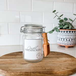 May include: A clear glass jar with a white label that reads "Carpet Freshener". The jar has a wooden scoop attached to the lid. The jar is sitting on a wooden surface.