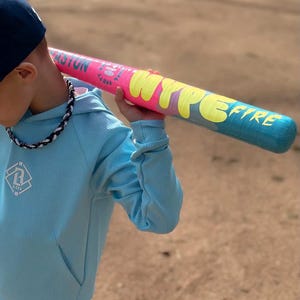 May include: A baseball bat with a pink and blue gradient design and the text "HYPE FIRE" in yellow. The person in the image is wearing a light blue hoodie and a dark blue baseball cap. The bat is a sporting good.