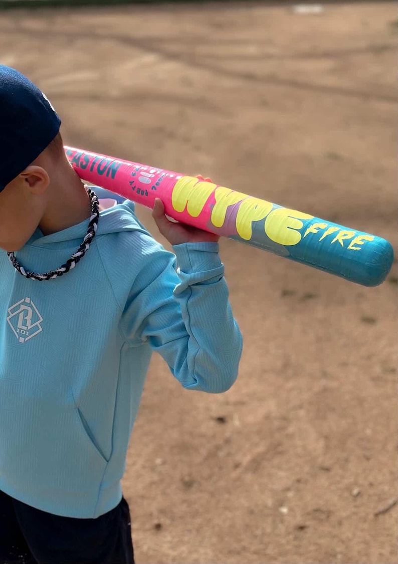 May include: A baseball bat with a pink and blue gradient design and the text "HYPE FIRE" in yellow. The person in the image is wearing a light blue hoodie and a dark blue baseball cap. The bat is a sporting good.