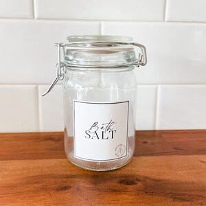 May include: A clear glass jar with a silver lid and a white label that says "Bath Salt".