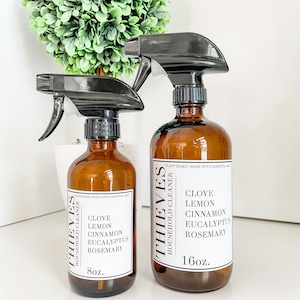 May include: Two amber glass spray bottles with black spray tops and white labels. The labels read "THIEVES HOUSEHOLD CLEANER" and list the ingredients: clove, lemon, cinnamon, eucalyptus, and rosemary. The smaller bottle is 8oz and the larger bottle is 16oz.