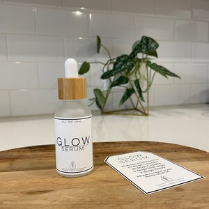 May include: A clear glass bottle with a bamboo lid and a white label that says "ALL NATURAL GLOW SERUM SKIN FREE LIVING". The bottle is sitting on a wooden surface with a white tag that says "GLOW SERUM 10 drops Frankincense 10 drops Lavender 10 drops Copaiba 5 drops Blue Tansy 2 drops Neroli Fill with rose hip oil or your favorite carrier oil SKIN FREE LIVING".