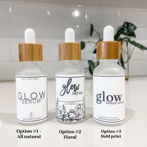 May include: Three glass bottles with dropper tops and bamboo lids. The bottles are labeled with different variations of the text "glow serum". The first bottle has the text "all natural" and "toxin free living". The second bottle has a floral design and the text "all natural made with essential oils". The third bottle has the text "beauty serum" and "made with essential oils".