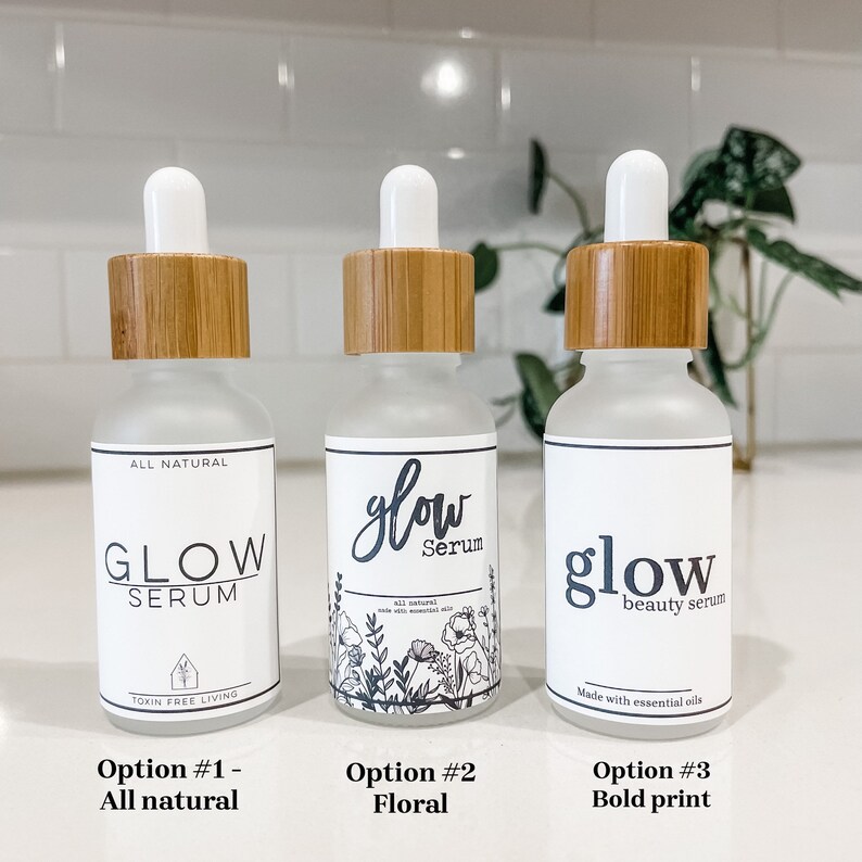 May include: Three glass bottles with dropper tops and bamboo lids. The bottles are labeled with different variations of the text "glow serum". The first bottle has the text "all natural" and "toxin free living". The second bottle has a floral design and the text "all natural made with essential oils". The third bottle has the text "beauty serum" and "made with essential oils".