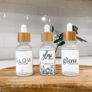 May include: Three glass bottles with bamboo lids and labels that say "Glow Serum". The bottles are on a wooden surface. The labels have different designs and text. One label says "All Natural" and "toxin free living". Another label says "Made with essential oils". The third label has a floral design and says "100% natural" and "made with essential oils".