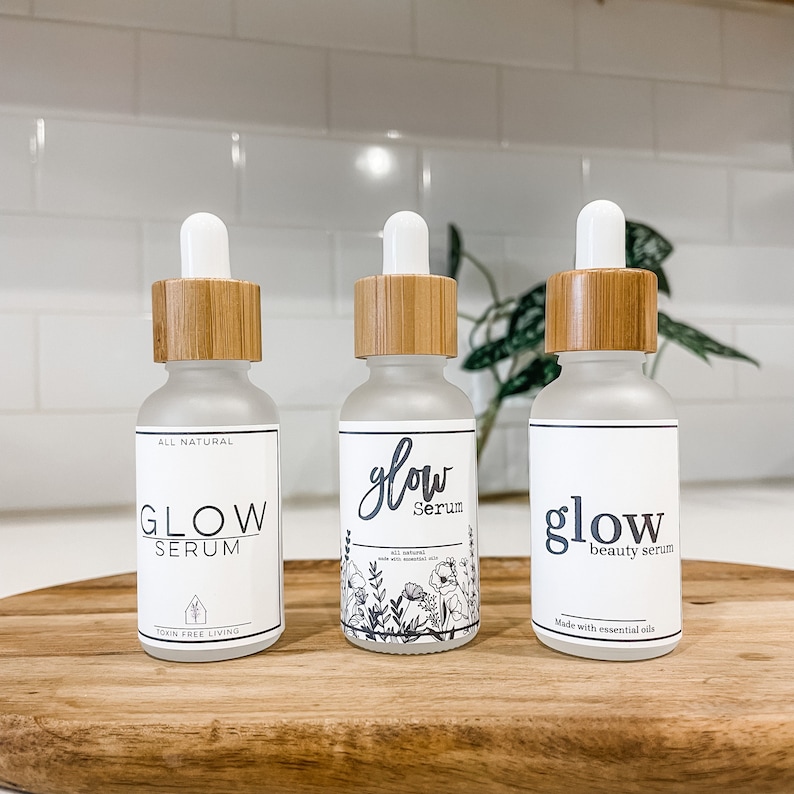 May include: Three glass bottles with bamboo lids and labels that say "Glow Serum". The bottles are on a wooden surface. The labels have different designs and text. One label says "All Natural" and "toxin free living". Another label says "Made with essential oils". The third label has a floral design and says "100% natural" and "made with essential oils".