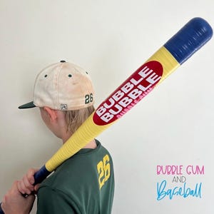 May include: A yellow baseball bat with blue ends and a red oval logo featuring the white text BUBBLE BUBBLE, paired with a cream-colored baseball cap with a green brim and the number 26, and a green shirt with the yellow number 26 on the back.