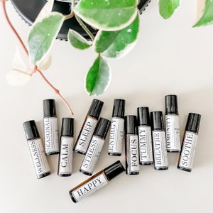 May include: A collection of ten small brown glass roller bottles with white labels. The labels have different words on them, including "Wellness", "Seasonal", "Calm", "Stress", "Sleep", "Energy", "Focus", "Tummy", "Breathe", "Immunity", and "Soothe".