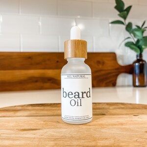 May include: A clear glass bottle with a bamboo lid and a white label that says "ALL NATURAL beard Oil MADE WITH ESSENTIAL OILS". The bottle is sitting on a wooden surface.