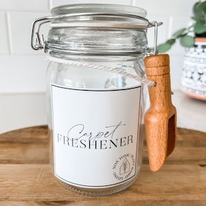 May include: A glass jar with a white label that says "Carpet Freshener" and a wooden scoop attached to the lid. The label also has a small circle with the text "Non Toxic Plant Based" in a circle.