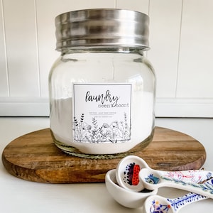 Puede incluir: Un frasco de vidrio con tapa plateada lleno de polvo blanco. El frasco tiene una etiqueta en blanco y negro que dice "laundry scentboost" con un diseño floral. La etiqueta también dice "no tóxico, limpieza a base de plantas, hecho con aceites esenciales".