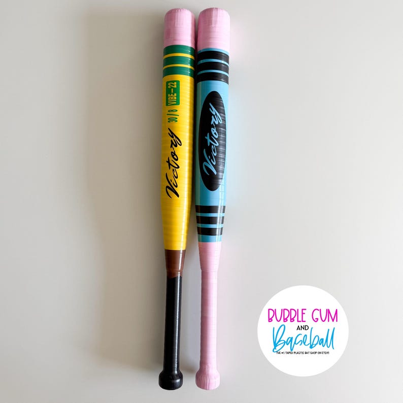 May include: Two plastic baseball bats with pencil-themed designs. The left bat is yellow with green stripes, brown and black handle, and text reading Victory 30/8 VIBE-22. The right bat is light blue with black stripes and text reading Victory.