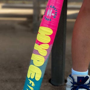 May include: A pink and blue baseball bat with the word "HYPE" in yellow and "FIRE" in yellow flames. The bat also has the text "EDITION" and "7003" in white. The bat is held near a person's leg and foot.