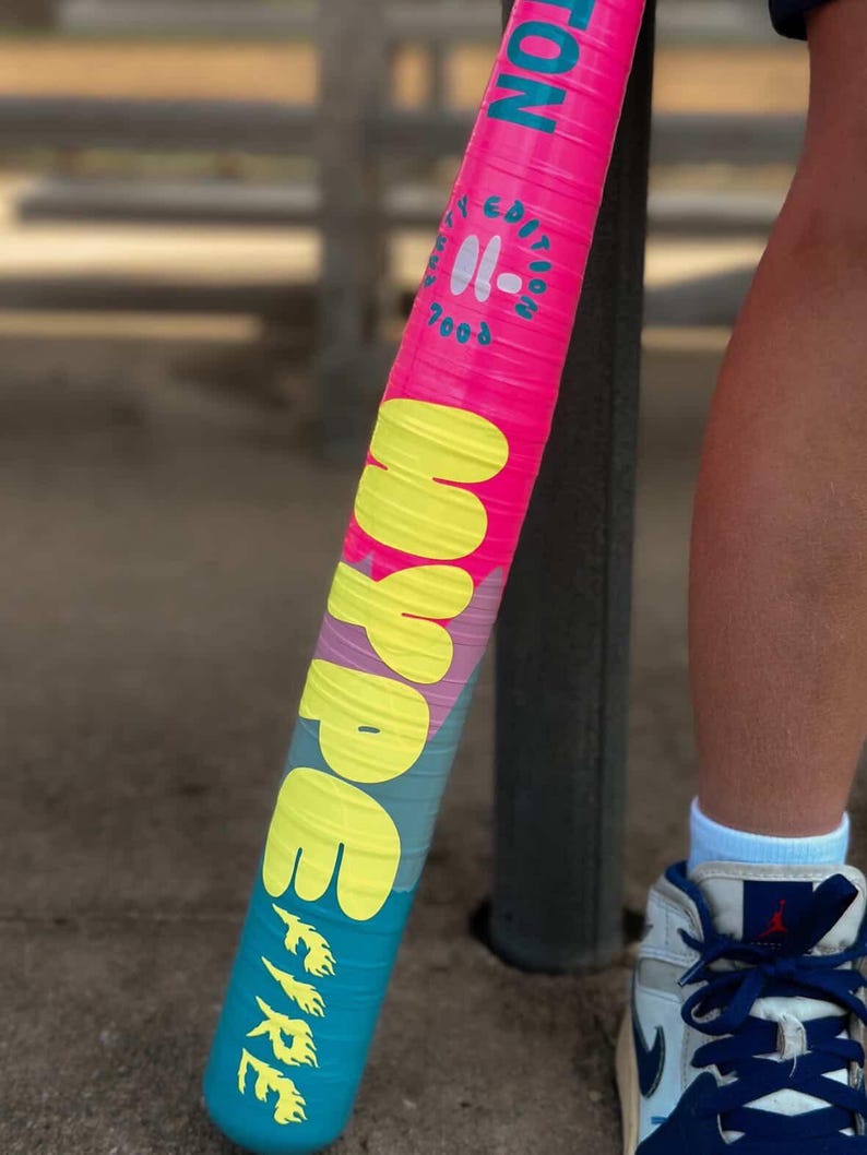 May include: A pink and blue baseball bat with the word "HYPE" in yellow and "FIRE" in yellow flames. The bat also has the text "EDITION" and "7003" in white. The bat is held near a person's leg and foot.