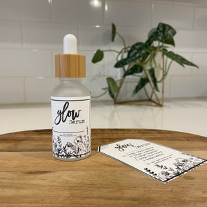 May include: A glass bottle with a bamboo lid and a white label that says "glow Serum" with a floral design. The bottle is sitting on a wooden surface with a white card that says "glow Serum" with instructions for use.