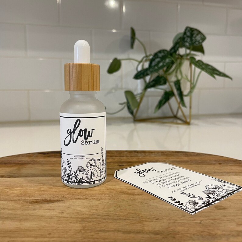 May include: A glass bottle with a bamboo lid and a white label that says "glow Serum" with a floral design. The bottle is sitting on a wooden surface with a white card that says "glow Serum" with instructions for use.