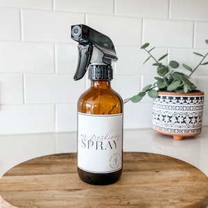 Könnte beinhalten: Eine braune Glasflasche mit einer schwarzen Sprühdüse, gefüllt mit Lufterfrischer. Die Flasche hat ein weißes Etikett mit dem Text "air freshener SPRAY" und ein kleines Design eines Hauses mit einem Baum.