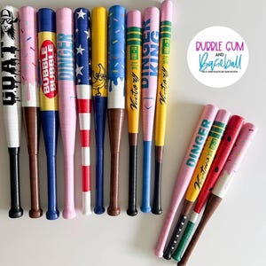 May include: A collection of colorful baseball bats with various designs and text. The bats feature patterns like stars, sprinkles, and stripes, along with the words "GOAT", "BUBBLE", "DINGER", "Victory", and "KING". The bats are in shades of pink, blue, yellow, and white.