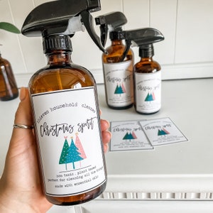 Op de afbeelding: Een bruine glazen spuitfles met een zwarte spuitmond. Het etiket luidt "thieves household cleaner Christmas spirit" met een feestelijk kerstboomontwerp. Het etiket vermeldt ook "niet giftig, plantaardig perfect voor het reinigen van alle dingen gemaakt met etherische oliën".