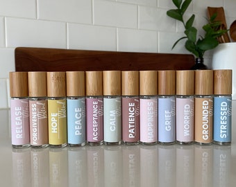 May include: A row of 12 small glass bottles with bamboo caps and labels. The labels are in pastel colors and have words like "Release", "Hope", "Peace", "Acceptance", "Calm", "Patience", "Happiness", "Grief", "Worried", "Grounded", and "Stressed".