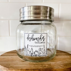 Op de afbeelding: Een glazen pot met een zilveren deksel, met het label "vaatwastabletten" met een bloemenmotief en de tekst "niet giftig, op plantenbasis reinigend gemaakt met etherische oliën".