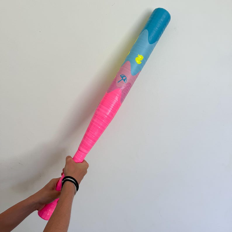 May include: A pink and blue baseball bat with a textured grip. The bat features a pink handle and a blue top, with a design of a yellow duck and a beach umbrella. The bat is held by a person.