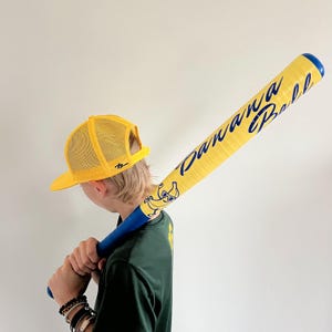 May include: A yellow baseball bat with the words "Banana Ball" in blue script. The bat features a cartoon banana character and a blue handle. A yellow mesh trucker hat is also visible.