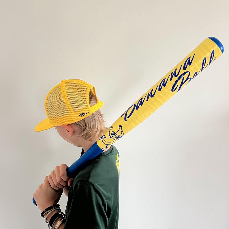 May include: A yellow baseball bat with the words "Banana Ball" in blue script. The bat features a cartoon banana character and a blue handle. A yellow mesh trucker hat is also visible.