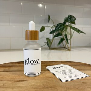 May include: A glass bottle with a bamboo lid and a white label that says "glow beauty serum" and "made with essential oils". A white card with the text "glow serum" and a list of ingredients is on a wooden surface.
