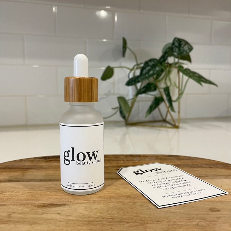 May include: A glass bottle with a bamboo lid and a white label that says "glow beauty serum" and "made with essential oils". A white card with the text "glow serum" and a list of ingredients is on a wooden surface.