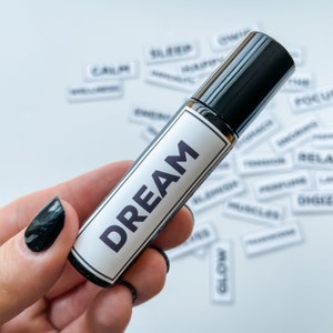 Pode incluir: Um frasco preto de rolo com um rótulo branco que diz "DREAM". O frasco é segurado em uma mão com esmalte de unha preto.