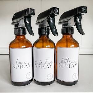 Op de afbeelding: Drie amberkleurige glazen spuitflessen met zwarte spuitkoppen en witte etiketten. De etiketten hebben zwarte tekst die "Linen Spray", "Air Freshener Spray" en "Bathroom Spray" luidt.