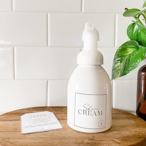 May include: White bottle with a pump dispenser labeled "Shave Cream". The bottle is sitting on a wooden surface with a small white label that says "Shave Cream" and "Forin Free Living".