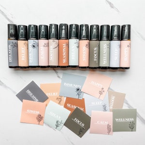 May include: A set of 12 essential oil roller bottles with labels for various uses, such as breathe, relief, immunity, calm, soothe, seasonal, relax, stress, focus, wellness, sleep, and happy. The bottles are arranged in two rows on a white marble surface. There are also 12 small cards with the same labels as the bottles, arranged in a similar pattern.