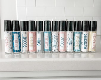 May include: A set of 14 small glass roller bottles with black caps and colorful labels. The labels have different words on them, including "Dream", "Believe", "Soothe", "Happy", "Calm", "Sweetness", "Wellness", "Focus", "Owie", "Tummy", "Butterflies", and "Immunity".