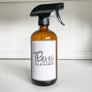 Może przedstawiać: Brązowa szklana butelka z rozpylaczem z czarną dźwignią. Butelka ma białą etykietę z tekstem "Thieves Household Cleaner".