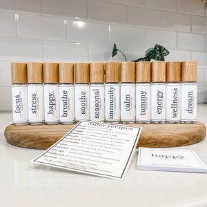 Può includere: Un set di 14 piccoli flaconi di vetro con rullo e tappi in bambù, ognuno etichettato con una parola diversa, come "focus", "stress", "happy", "breathe", "soothe", "seasonal", "immunity", "calm", "tummy", "energy", "wellness" e "dream". I flaconi sono disposti in fila su una superficie di legno. Una carta con le parole "roller recipes" è visibile in primo piano.