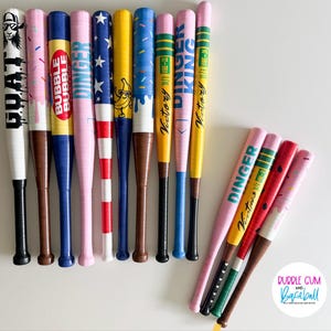 May include: A collection of colorful baseball bats with various designs and text. The bats feature a variety of colors, including pink, blue, yellow, and red, with graphics and text such as "GOAT", "BUBBLE", and "DINGER".