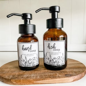 Puede incluir: Dos botellas de vidrio ámbar con tapas de bomba negras y etiquetas. La botella de la izquierda dice "jabón de manos" y la de la derecha dice "jabón para platos". Ambas etiquetas tienen un diseño floral y el texto "no tóxico, limpieza a base de plantas, hecho con aceites esenciales."