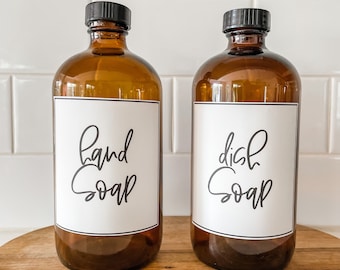 May include: Two amber glass bottles with black lids and white labels. The labels have black handwritten text that reads "hand soap" and "dish soap".
