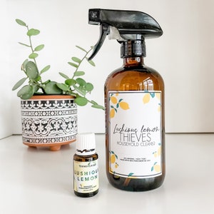 May include: A brown glass bottle with a black spray nozzle filled with a clear liquid. The bottle has a white label with the text "Lushious lemon THIEVES HOUSEHOLD CLEANER" and a lemon design. A small brown glass bottle with a white cap and a white label with the text "LUSHIOUS LEMON" is in front of the larger bottle.