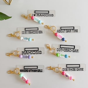 May include: Six rectangular acrylic keychains with gold-tone lobster clasps. Each features black text including "I love Mahjong", "In My Mahjong Era", "About Bam Time", "Mahjong", "Dot Crack Bam", "Mahjong With Mah Girls", and "One More Game".